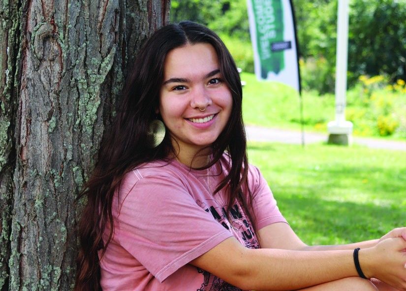 A young woman with long dark hair sits outdoors, leaning against a tree. She is smiling at the camera, wearing a pink T-shirt, large circular earrings, and a nose ring. The background shows a grassy area with trees and a blurred banner in the distance. The scene is bright and peaceful.