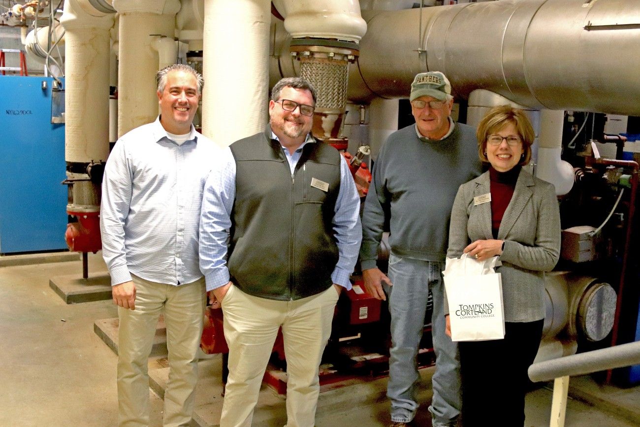 A group of four people is standing in front of industrial machinery. The individuals are smiling, casually dressed, and appear to be on a tour or working in a technical environment. Two men on the left are wearing light-colored shirts, with one wearing a vest. The third man, standing next to them, is wearing a cap and a sweater. The woman on the right, holding a bag labeled "Tompkins Cortland Community College," is dressed professionally with glasses and a name tag. The background consists of large pipes an