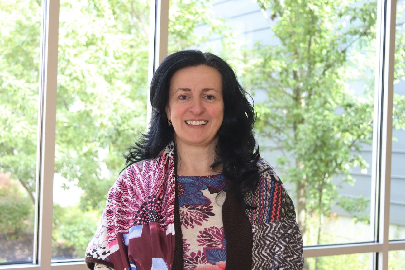 Woman with long black hair smiling in front of a large window with greenery visible outside. She is wearing a colorful, patterned outfit that includes floral and geometric designs, with a mix of red, purple, and blue colors.