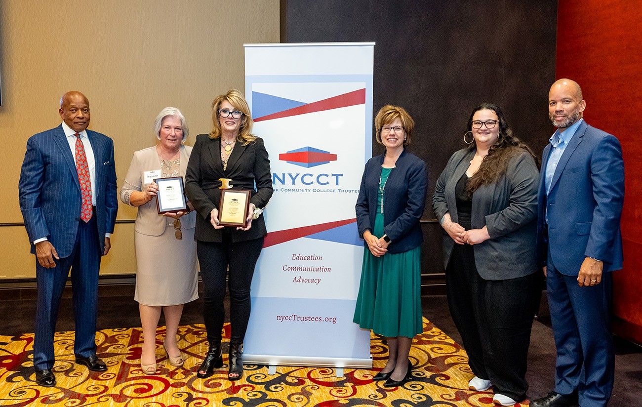 Group photo of six individuals standing together in front of a New York Community College Trustees (NYCCT) banner. Two women in the center hold plaques, presumably awards, while the others stand on either side, dressed in professional attire. The background has a beige and dark accent wall, and the floor is decorated with a patterned carpet.