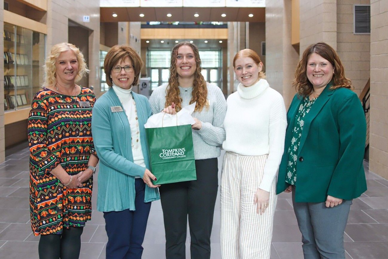 Five individuals standing in a brightly lit hallway, posing for a group photo. The central figure holds a green Tompkins Cortland Community College gift bag, smiling warmly. The others, dressed in professional attire, stand on either side, also smiling.