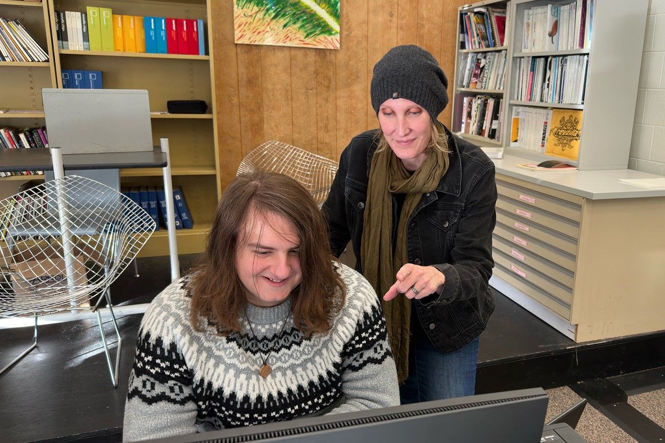 A woman wearing a beanie and scarf stands next to a young man with long hair, who is seated and smiling at a computer screen. The woman is pointing at the screen, engaging with him. They are in a room with bookshelves, colorful folders, and art on the wall.