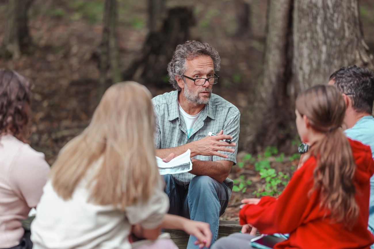 A bearded man wearing glasses and a plaid shirt, holding a pen and some papers, sits outdoors in a wooded area, engaged in a discussion with a small group of students. The students, seated around him, appear attentive, with notebooks in hand. The natural, forested background adds a sense of immersion in nature to the learning setting.