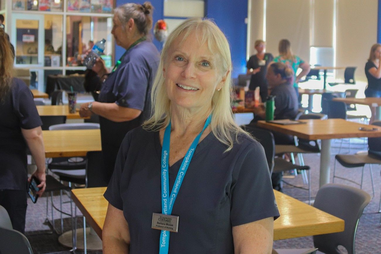 A smiling woman with blonde hair, wearing a Tompkins Cortland Community College lanyard and a gray scrub top, stands in a well-lit room filled with tables and people. She appears confident and approachable, with a name tag that reads "Barbara Moose, Professor of Nursing." Other individuals are visible in the background, engaging in conversation and activities.