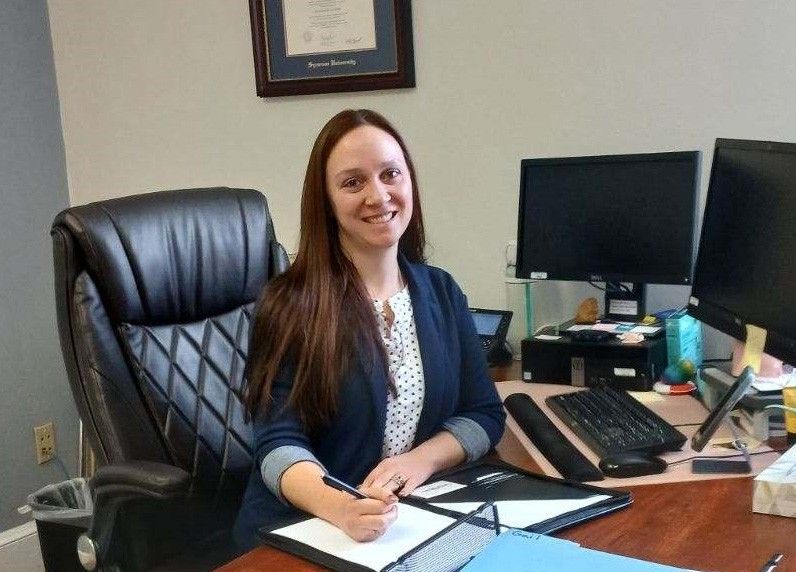  A professional woman with long brown hair is seated at a desk in an office. She is wearing a navy blazer over a white patterned blouse and is smiling while holding a pen and writing in a folder. The desk is equipped with dual computer monitors, a keyboard, and various office supplies. A framed certificate hangs on the wall behind her, and a black office chair is visible in the foreground.