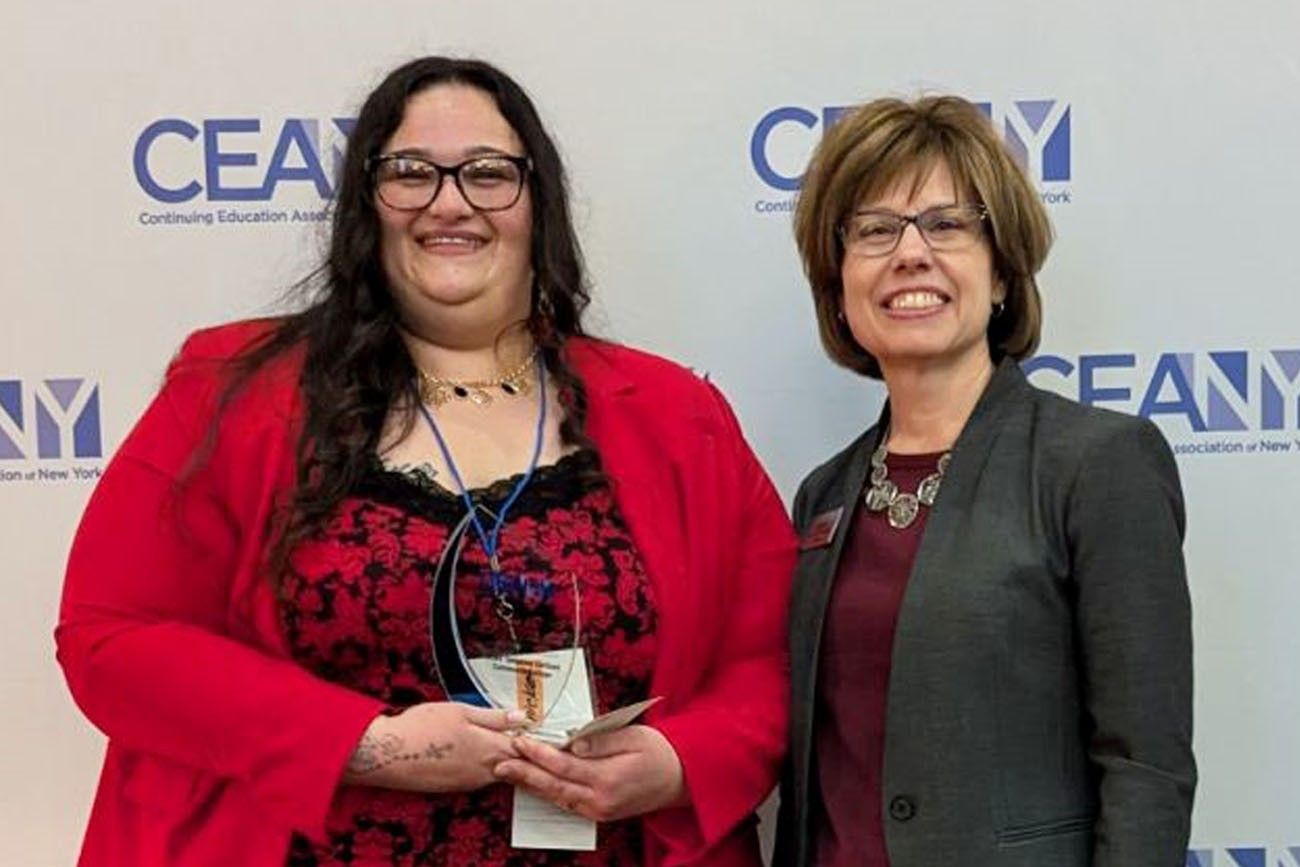 Two women standing together at a CEANY (Continuing Education Association of New York) event. The woman on the left has long dark hair, glasses, and is wearing a red jacket over a black and red lace top, holding an award. The woman on the right has short brown hair, glasses, and is wearing a gray blazer over a maroon top, smiling.