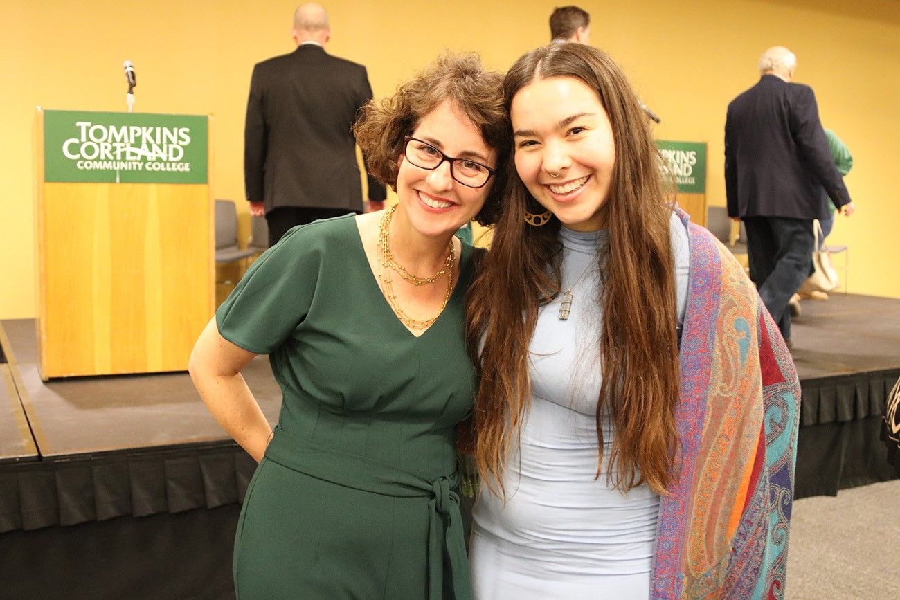 Two smiling women pose together in front of a stage at an event. The woman on the left is wearing glasses and a green dress, accessorized with a gold necklace. The woman on the right is wearing a light blue dress with a colorful patterned shawl. Behind them is a podium with a sign reading "Tompkins Cortland Community College" and several people walking on or near the stage.