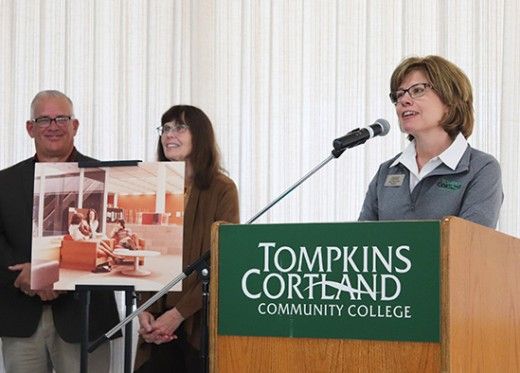 Three individuals stand at a podium during an event at Tompkins Cortland Community College. A woman at the podium is speaking into a microphone, wearing glasses and a name tag, while a green sign on the podium displays the college's name. Behind her, a man and another woman hold a large image of what appears to be a design or architectural concept. The atmosphere is formal.