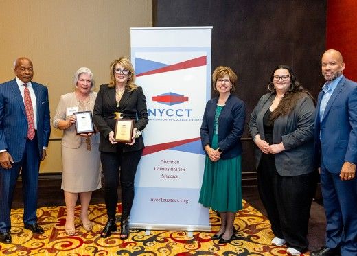 Group photo of six individuals standing together in front of a New York Community College Trustees (NYCCT) banner. Two women in the center hold plaques, presumably awards, while the others stand on either side, dressed in professional attire. The background has a beige and dark accent wall, and the floor is decorated with a patterned carpet.