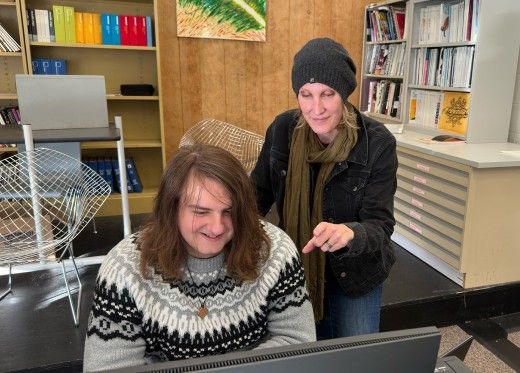 A woman wearing a beanie and scarf stands next to a young man with long hair, who is seated and smiling at a computer screen. The woman is pointing at the screen, engaging with him. They are in a room with bookshelves, colorful folders, and art on the wall.