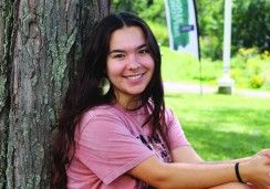 A young woman with long dark hair sits outdoors, leaning against a tree. She is smiling at the camera, wearing a pink T-shirt, large circular earrings, and a nose ring. The background shows a grassy area with trees and a blurred banner in the distance. The scene is bright and peaceful.