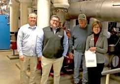 A group of four people is standing in front of industrial machinery. The individuals are smiling, casually dressed, and appear to be on a tour or working in a technical environment. Two men on the left are wearing light-colored shirts, with one wearing a vest. The third man, standing next to them, is wearing a cap and a sweater. The woman on the right, holding a bag labeled "Tompkins Cortland Community College," is dressed professionally with glasses and a name tag. The background consists of large pipes an