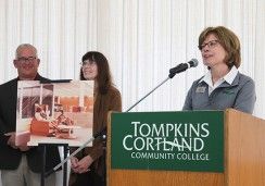 Three individuals stand at a podium during an event at Tompkins Cortland Community College. A woman at the podium is speaking into a microphone, wearing glasses and a name tag, while a green sign on the podium displays the college's name. Behind her, a man and another woman hold a large image of what appears to be a design or architectural concept. The atmosphere is formal.