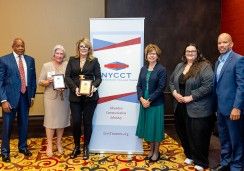 Group photo of six individuals standing together in front of a New York Community College Trustees (NYCCT) banner. Two women in the center hold plaques, presumably awards, while the others stand on either side, dressed in professional attire. The background has a beige and dark accent wall, and the floor is decorated with a patterned carpet.