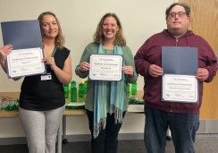 Three people are standing side by side, each holding a "Certificate of Achievement." From left to right, the certificates read: "Angela Carl," "Mandy Darling," and "William Henry Vaughn-Russell." All three are smiling, standing in front of a table with green water bottles and festive decorations in the background.