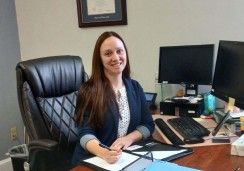  A professional woman with long brown hair is seated at a desk in an office. She is wearing a navy blazer over a white patterned blouse and is smiling while holding a pen and writing in a folder. The desk is equipped with dual computer monitors, a keyboard, and various office supplies. A framed certificate hangs on the wall behind her, and a black office chair is visible in the foreground.