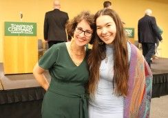 Two smiling women pose together in front of a stage at an event. The woman on the left is wearing glasses and a green dress, accessorized with a gold necklace. The woman on the right is wearing a light blue dress with a colorful patterned shawl. Behind them is a podium with a sign reading "Tompkins Cortland Community College" and several people walking on or near the stage.